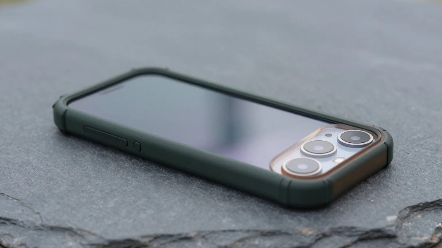 Smartphone in dunkelgrüner Schutzhülle mit erhöhtem Rand liegt mit der Rückseite nach oben auf Schiefer, daneben eine bernsteinfarbene Hülle