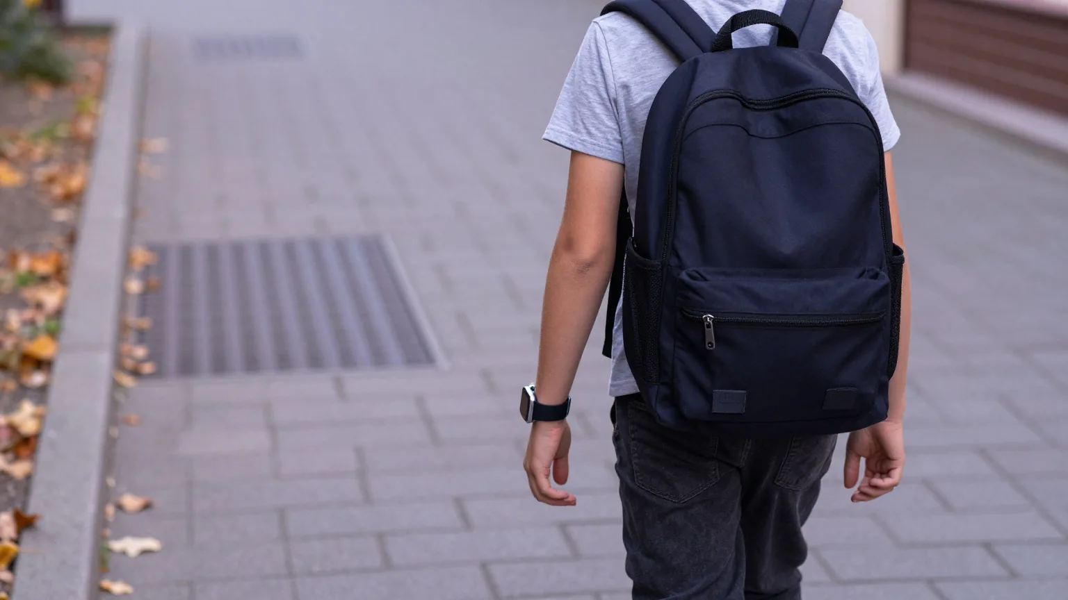 Junger Mensch von hinten mit Schulrucksack auf einem Gehweg einer Kleinstadt, am Handgelenk eine Smartwatch mit dunklem Display