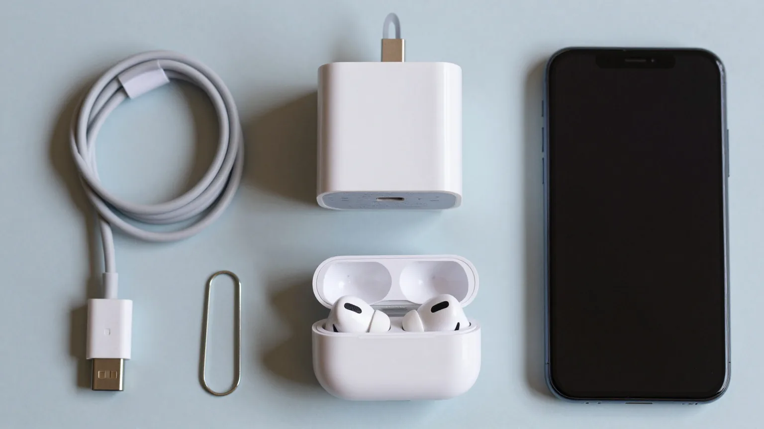 Von oben fotografiert: USB-C-Kabel, Ladegerät, Ohrhörer, SIM-Nadel und ein Smartphone ordentlich im Raster auf hellblauem Grund