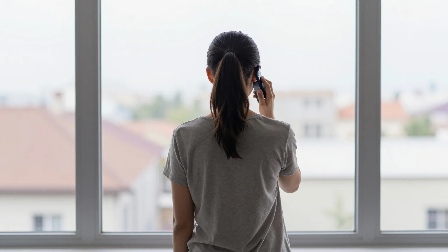 Frau steht von hinten gesehen an einem großen Fenster und telefoniert