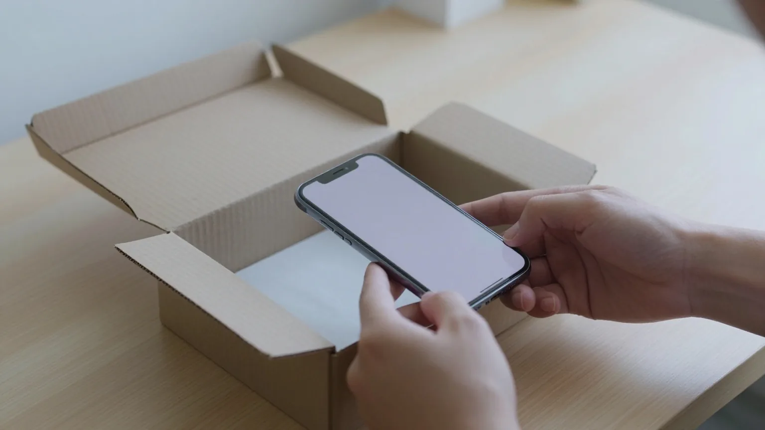 Zwei Hände legen ein Smartphone mit der Rückseite nach oben in einen Versandkarton mit Packpapier