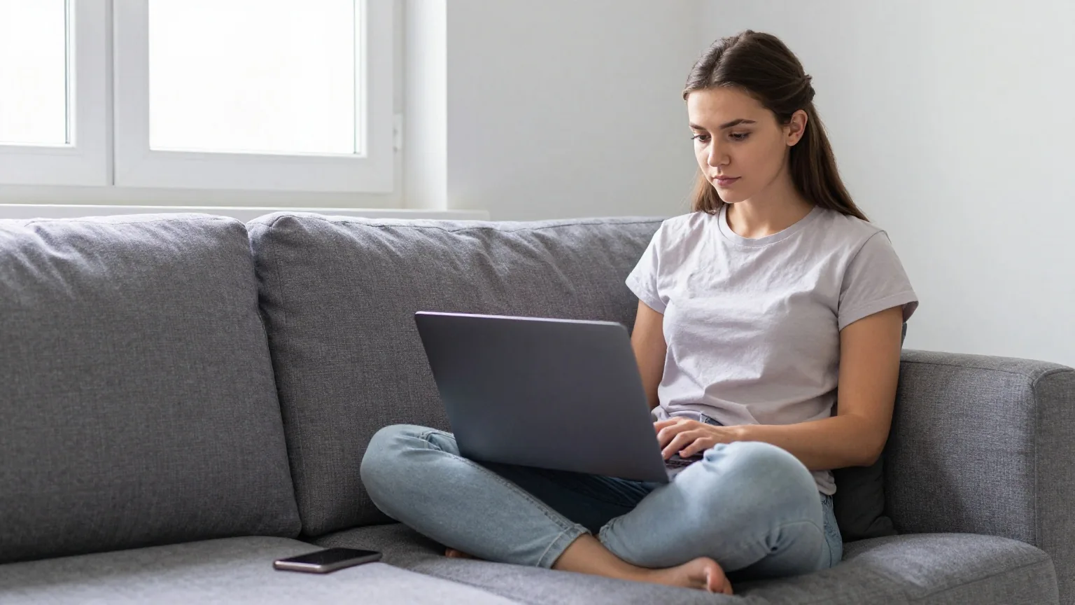 Junge Frau im Schneidersitz auf dem Sofa, auf den Knien ein Laptop mit abgewandtem Bildschirm, neben ihr ein umgedrehtes Smartphone
