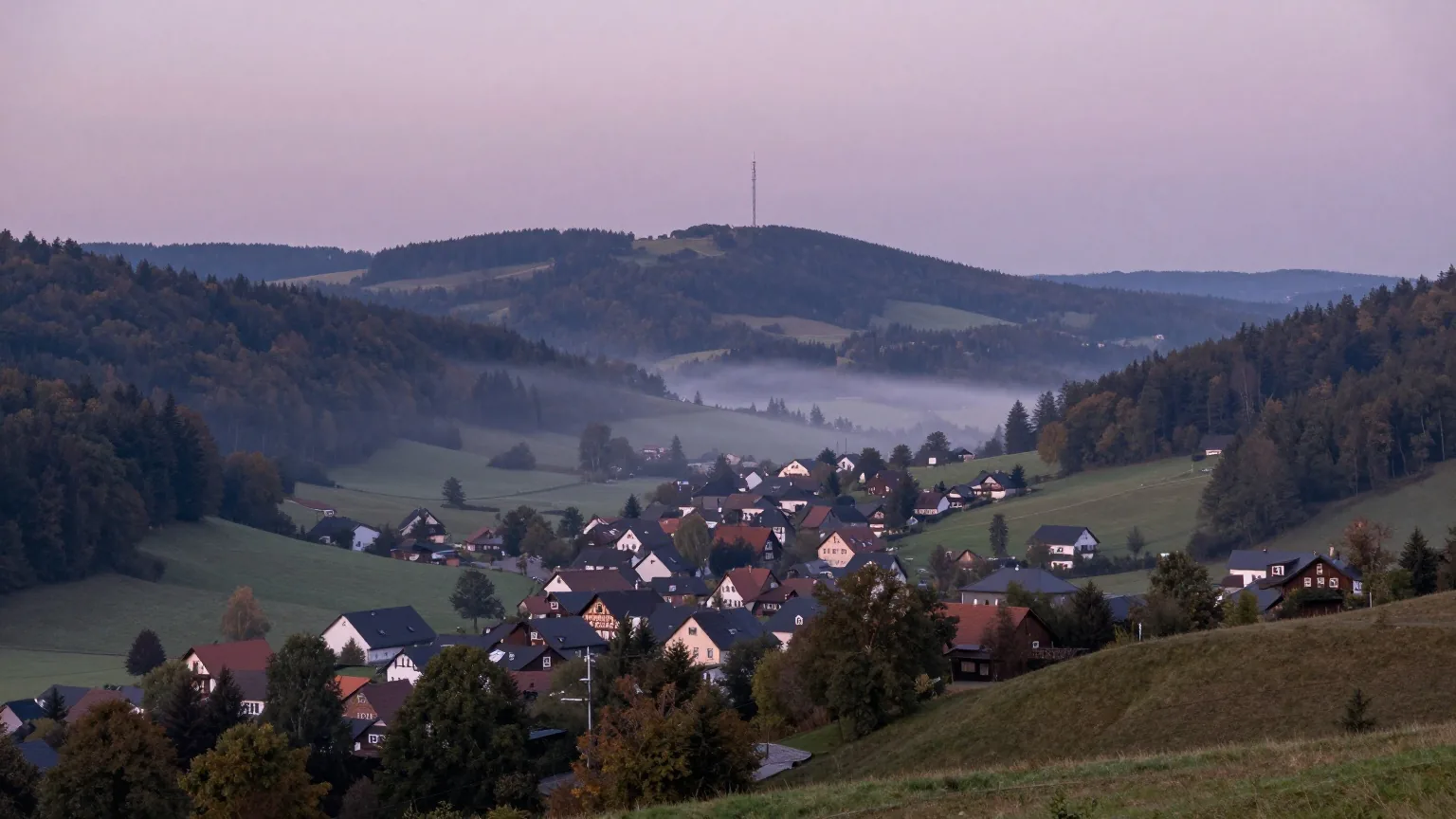 Dorf in einem engen, bewaldeten Tal in der Dämmerung, auf einem fernen Bergkamm ein kaum sichtbarer Mobilfunkmast