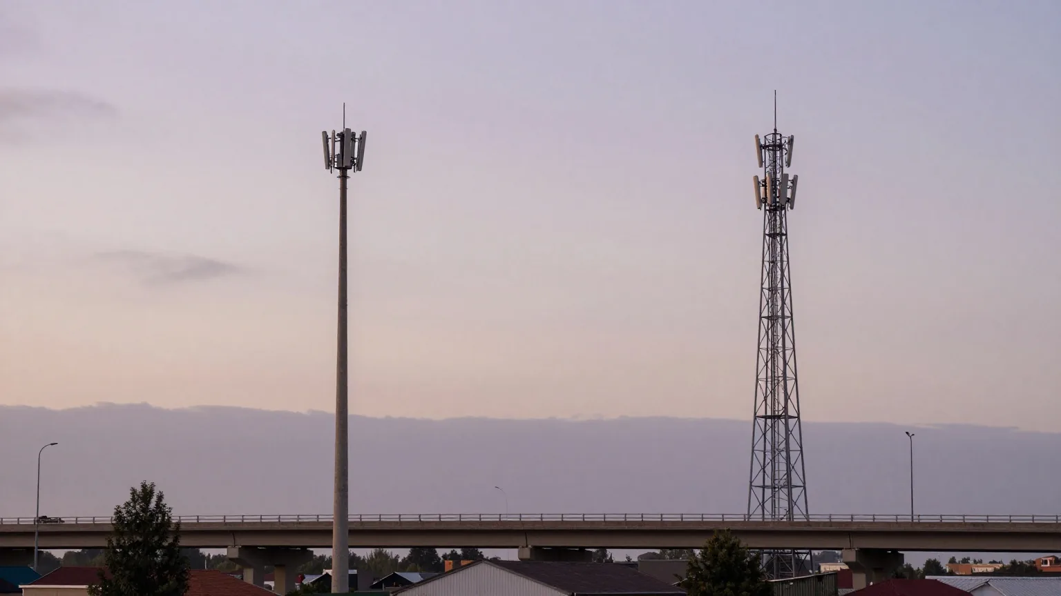 Zwei unterschiedliche Mobilfunkmasten am Rand einer Kleinstadt, dazwischen eine Autobahnbrücke unter bewölktem Abendhimmel