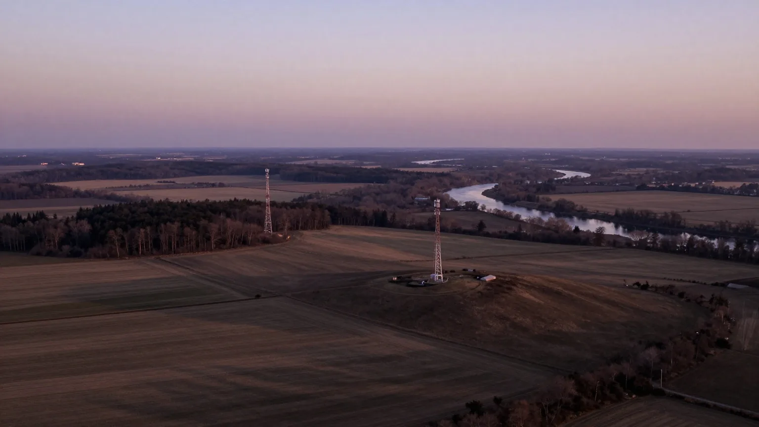 Luftbild einer Landschaft aus Feldern, Wald und Flusstal in der Dämmerung, auf Hügeln stehen weit verteilt drei Mobilfunkmasten