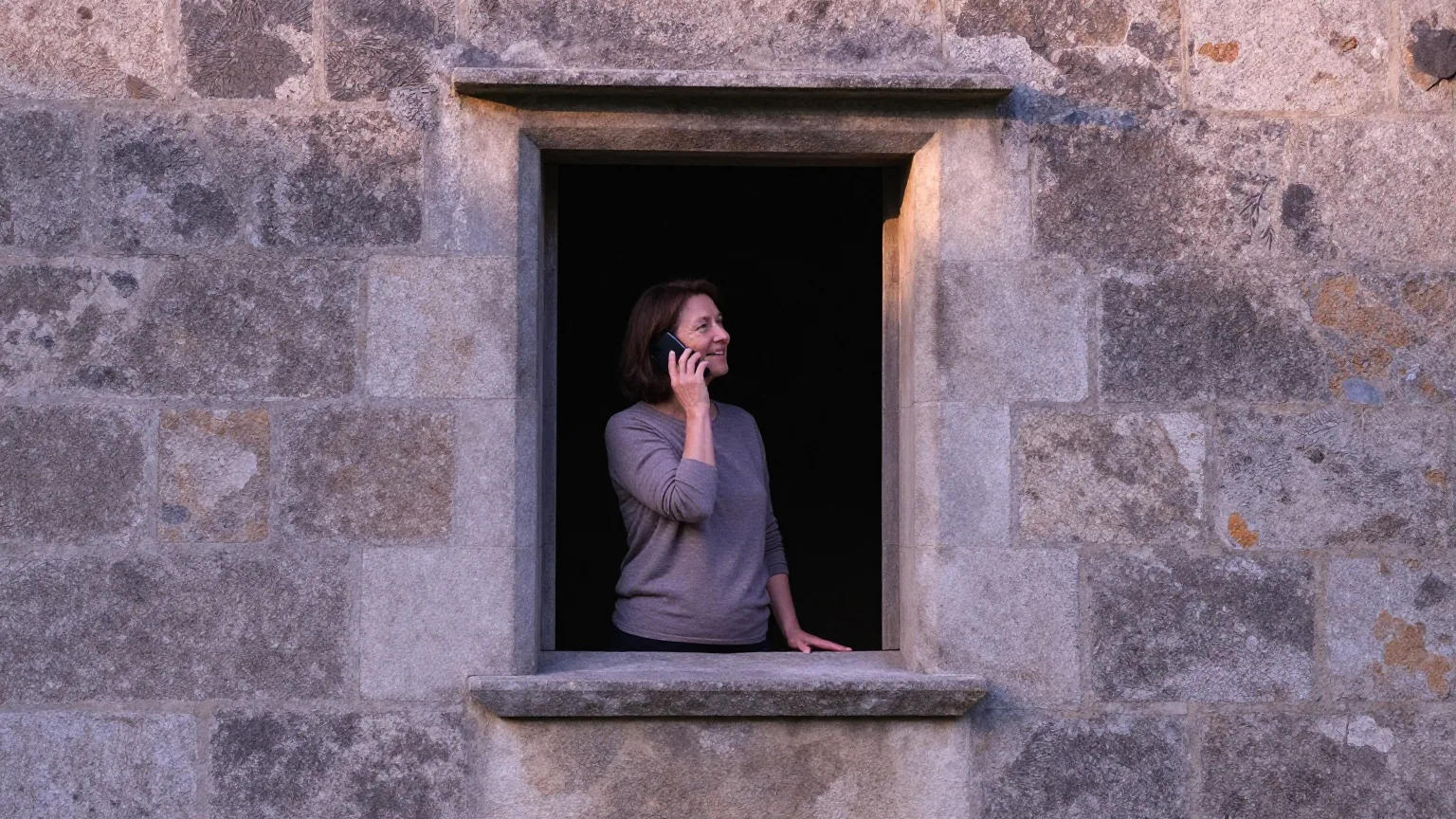 Frau steht in der Fensternische eines alten Steinhauses und telefoniert, von der Seite gesehen