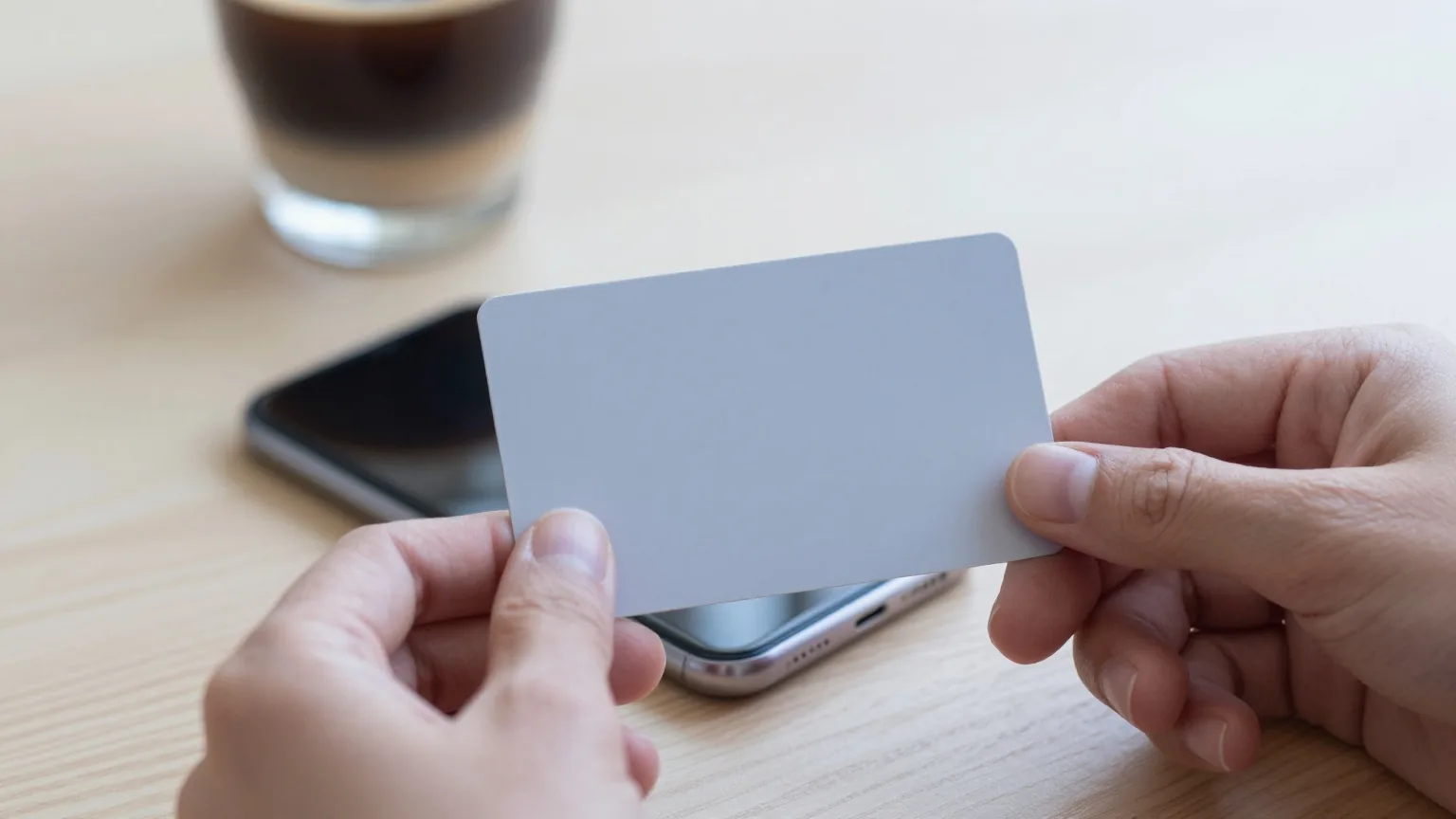 Hände halten eine unbedruckte Plastikkarte flach an die Rückseite eines Smartphones über einem hellen Holztisch