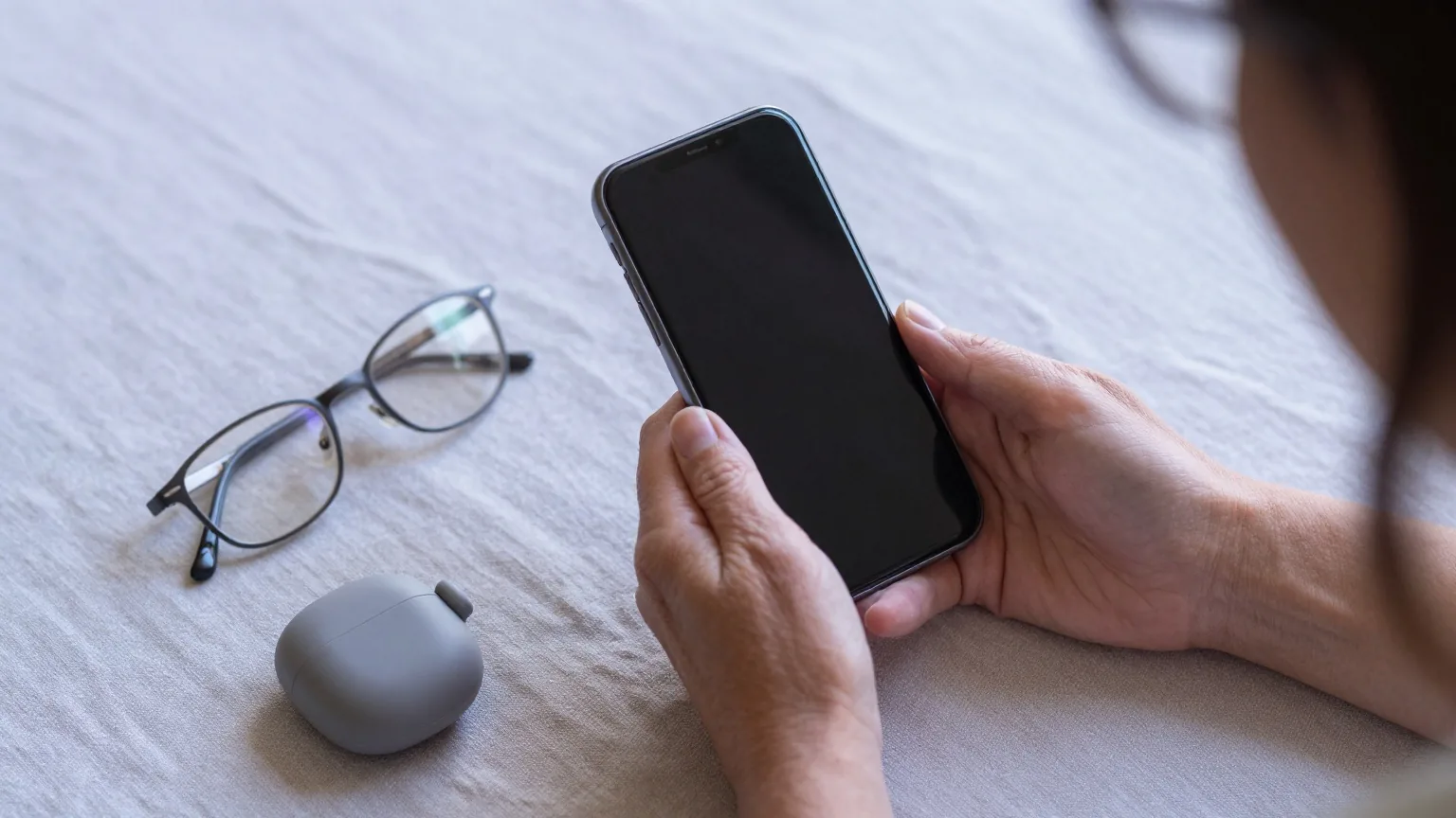 Hände einer älteren Frau halten ein Smartphone mit dunklem Display, daneben liegen eine Lesebrille und ein Etui für Hörgeräte