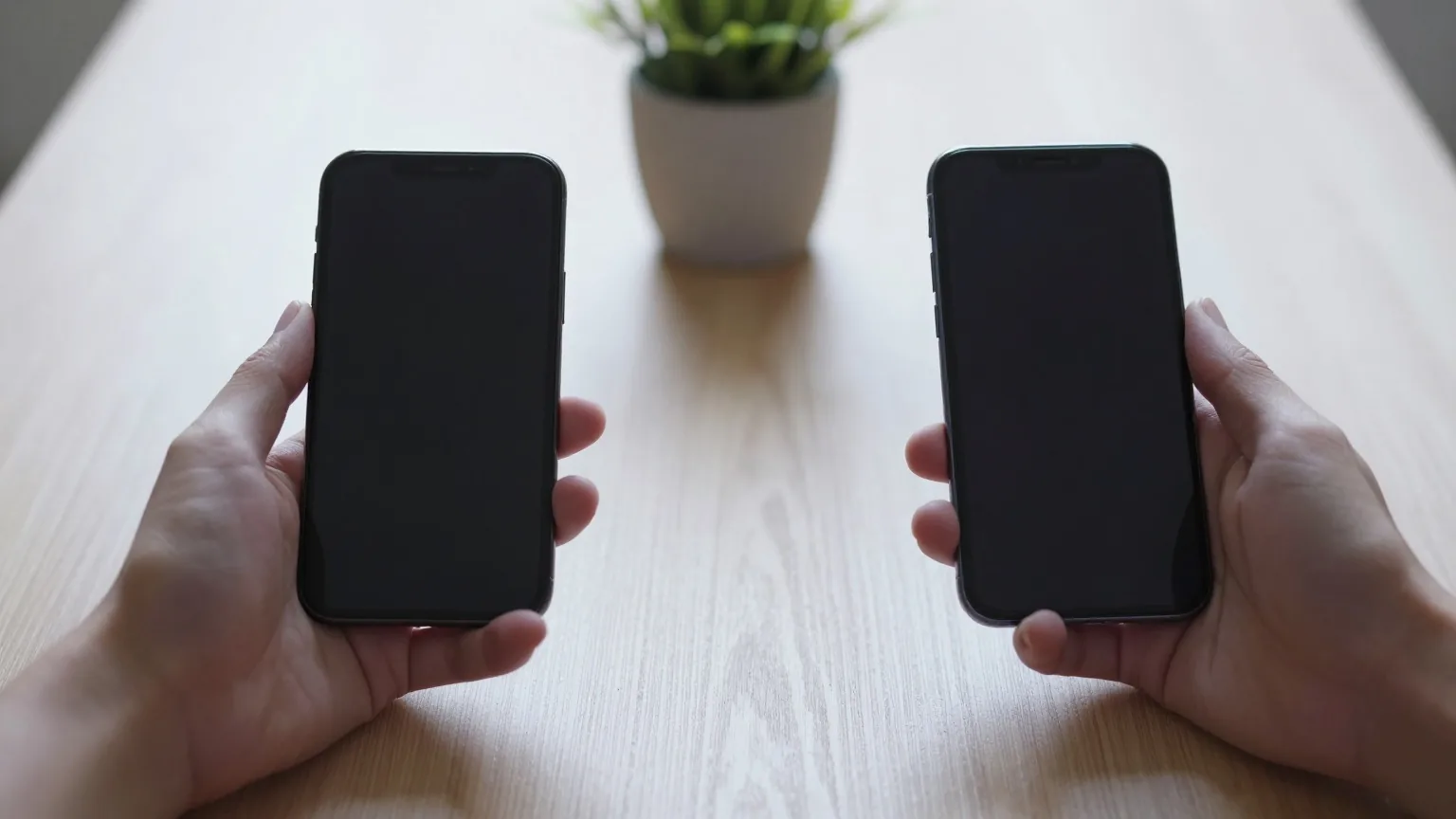 Zwei Hände halten zwei Smartphones mit dunklem Display nebeneinander über einen hellen Holztisch