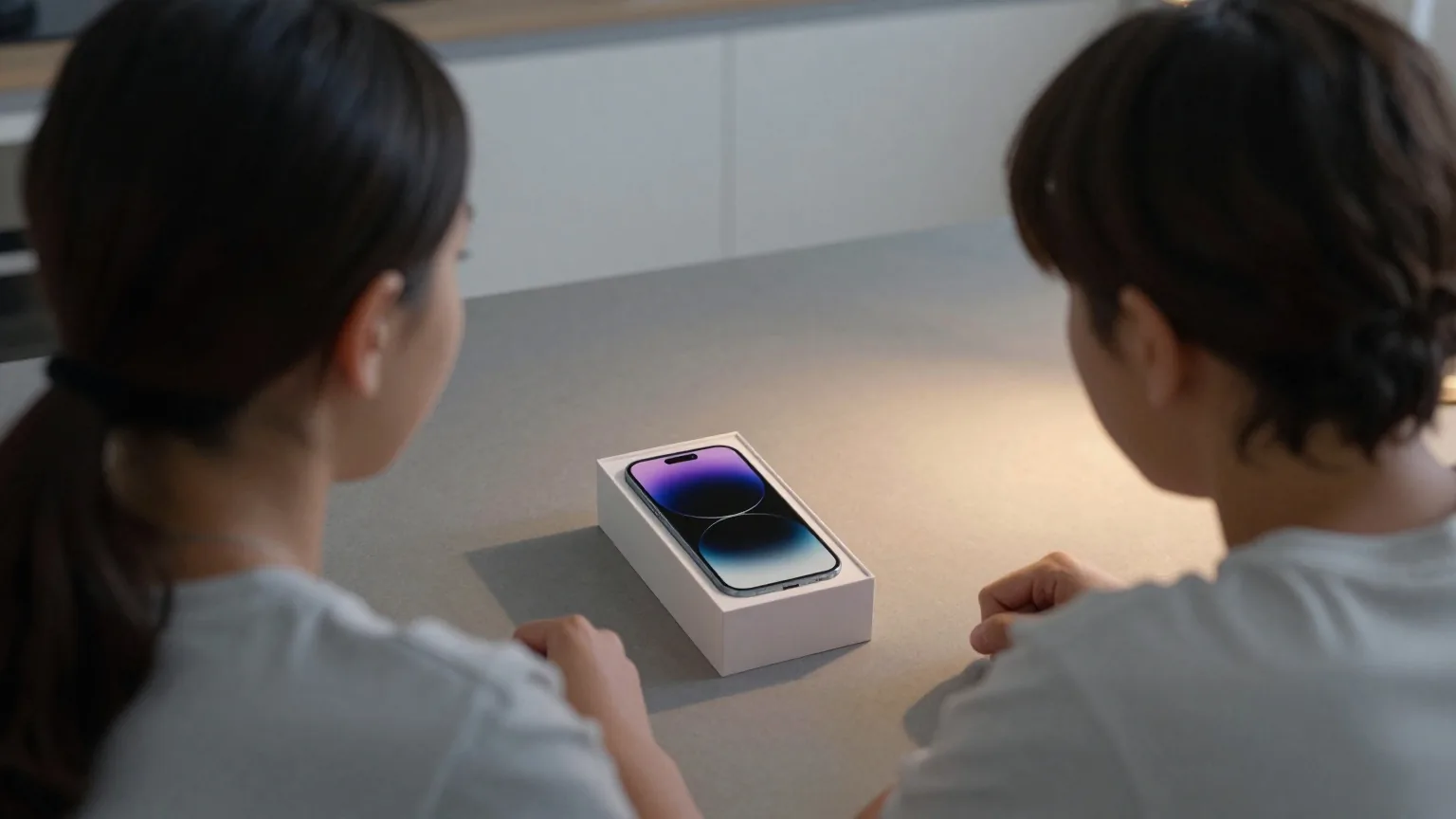 Mutter und junger Mensch von hinten am Küchentisch, zwischen ihnen ein neues Smartphone mit dunklem Display neben seinem Karton