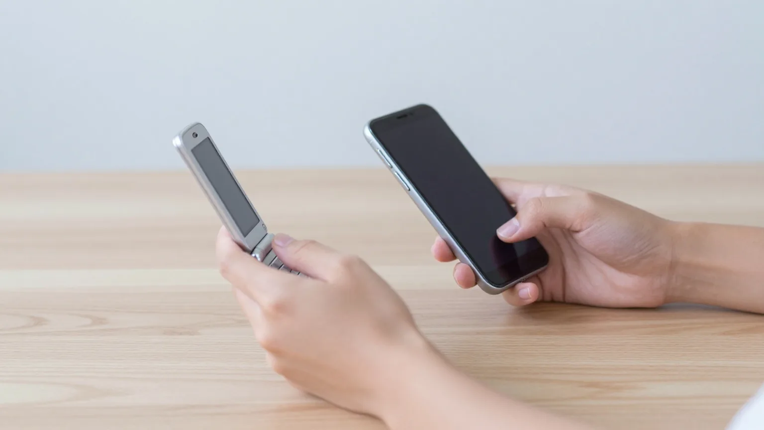 Zwei Hände über einem hellen Holztisch, eine hält ein geschlossenes silbernes Klapphandy, die andere ein Smartphone mit dunklem Display