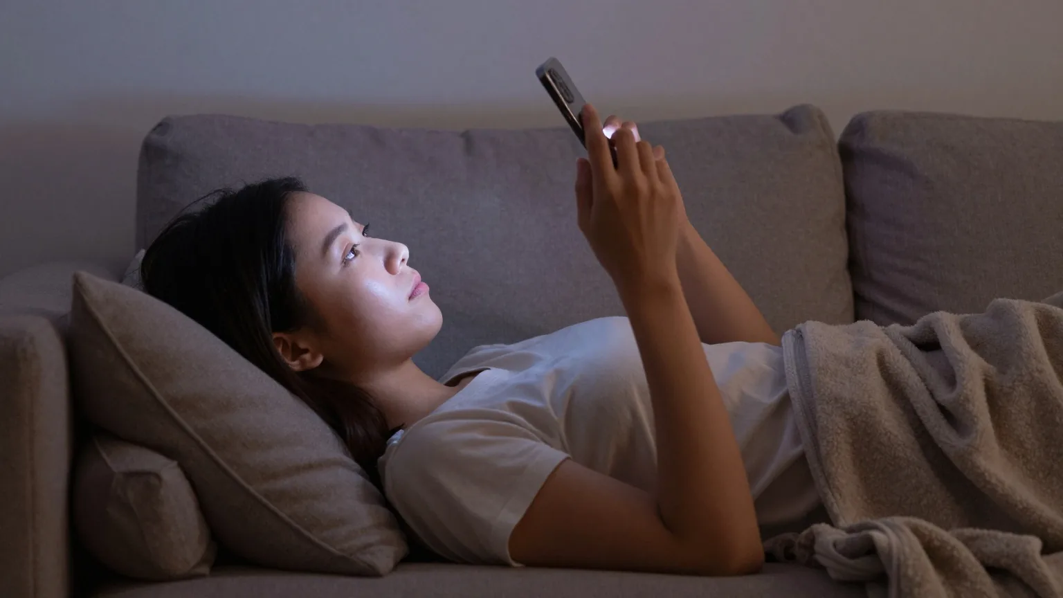 Junge Frau liegt abends von der Seite gesehen auf einem Sofa und hält ein Smartphone mit der Rückseite zur Kamera über sich