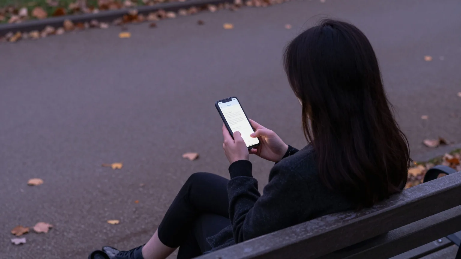 Frau sitzt von hinten gesehen in der Dämmerung auf einer Parkbank und hält ein Smartphone, dessen Display abgewandt ist