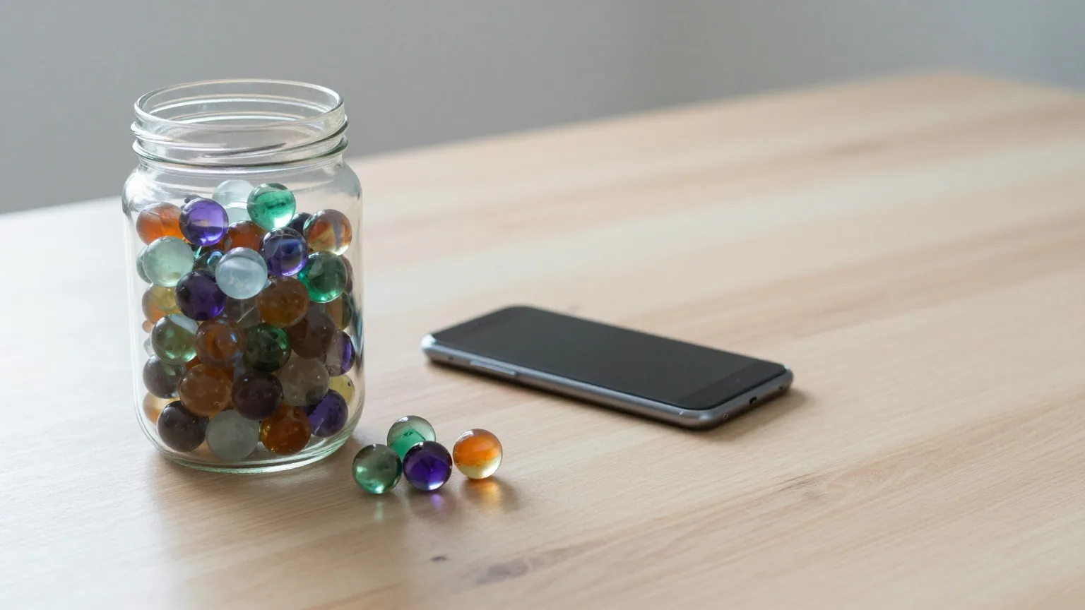 Großes Glas, halb gefüllt mit bunten Murmeln, auf einem hellen Eichentisch, daneben ein Smartphone mit der Rückseite nach oben