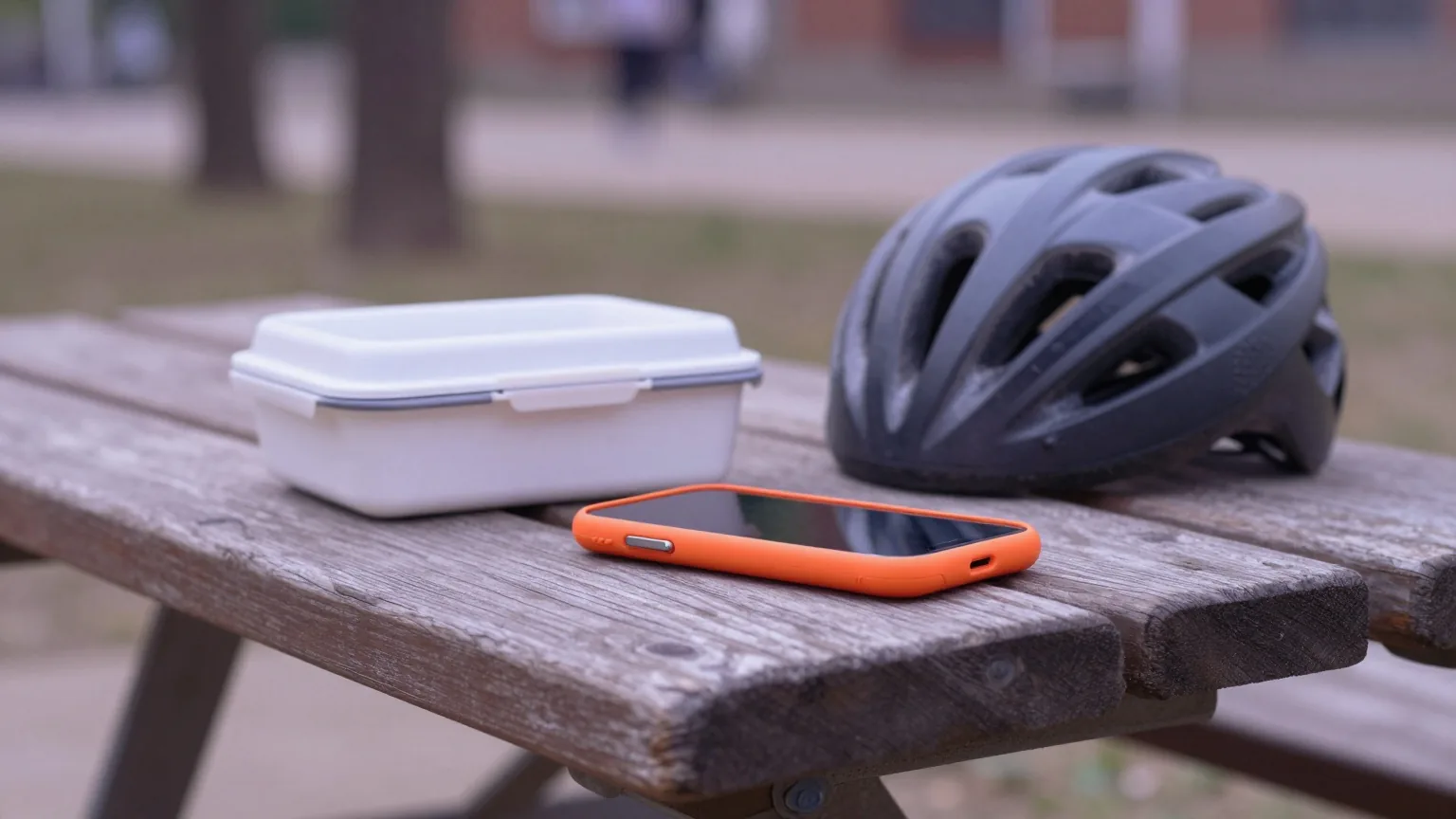 Smartphone in orangefarbener Hülle liegt mit dem Display nach unten auf einer Picknickbank im Schulhof neben Brotdose und Fahrradhelm