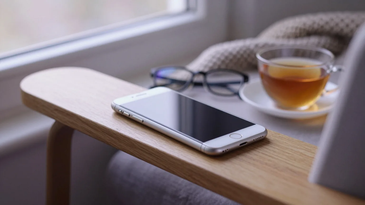 Smartphone liegt mit der Rückseite nach oben auf der Armlehne eines Lesesessels, daneben Lesebrille und eine Tasse Tee