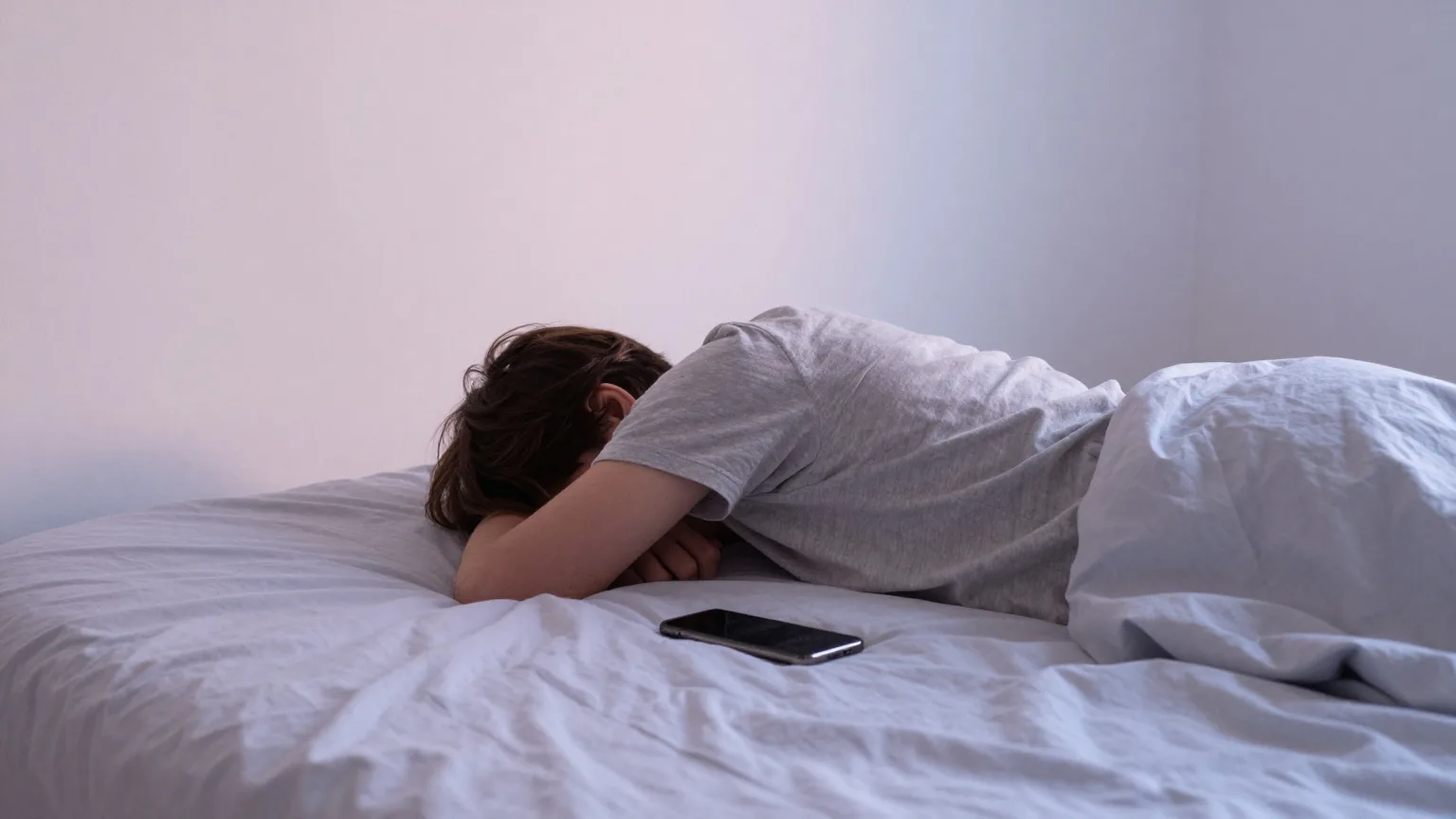 Junger Mensch von hinten auf einem Bett liegend, neben ihm ein umgedrehtes Smartphone auf der hellen Bettdecke